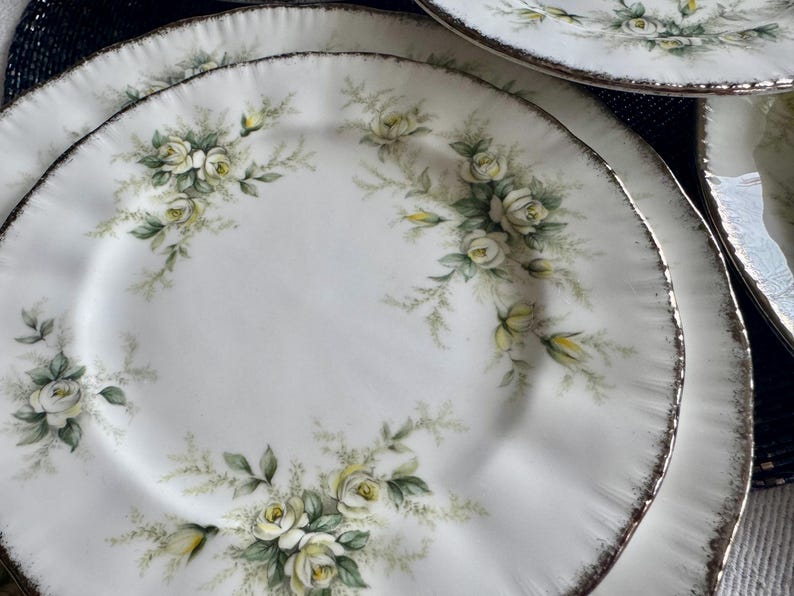 May include: A set of white ceramic plates with a delicate floral design featuring white roses and green leaves. The plates have a silver rim.