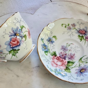 Pu&ograve; includere: Un set vintage di tazza e piattino da t&egrave; con disegni floreali rosa, blu e viola. La tazza e il piattino hanno bordi dorati e sono posti su un vassoio di marmo bianco con manici dorati. La tazza da t&egrave; &egrave; inclinata.