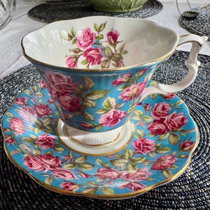 K&ouml;nnte beinhalten: Ein blaues und wei&szlig;es Tee-Set mit einem Blumenmuster aus rosa Rosen. Tasse und Untertasse haben einen goldenen Rand.