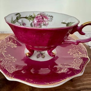 May include: A vintage teacup and saucer set. The cup and saucer are a deep magenta color with gold trim. The cup features a floral design inside, and the saucer has a gold leaf pattern. The cup has a handle with gold accents.