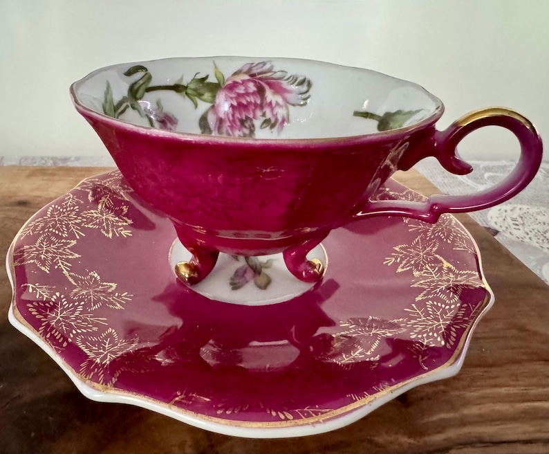 May include: A vintage teacup and saucer set. The cup and saucer are a deep magenta color with gold trim. The cup features a floral design inside, and the saucer has a gold leaf pattern. The cup has a handle with gold accents.