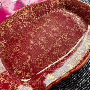 May include: A red and gold serving platter with a floral pattern. The platter has a scalloped edge and is made of ceramic.