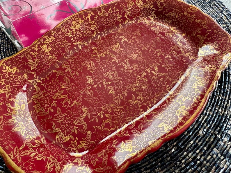 May include: A red and gold serving platter with a floral pattern. The platter has a scalloped edge and is made of ceramic.