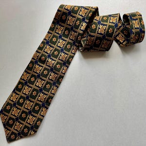 May include: A necktie featuring a dark green background with a repeating grid pattern of gold-colored floral motifs and small dark blue diamond shapes. The tie is laid out flat with the top portion folded over itself in a loose coil.