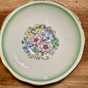 May include: A white teacup saucer with a green rim and gold trim. The saucer features a floral design with pink, yellow, and blue flowers.