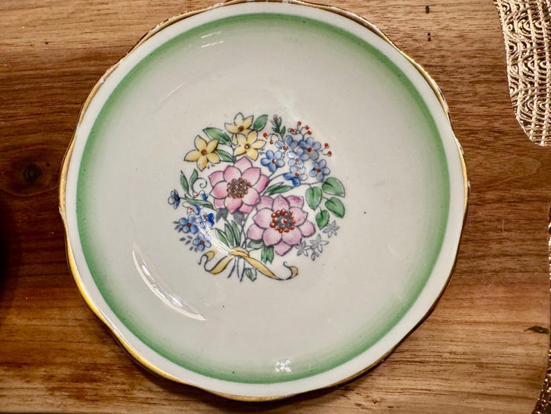 May include: A white teacup saucer with a green rim and gold trim. The saucer features a floral design with pink, yellow, and blue flowers.
