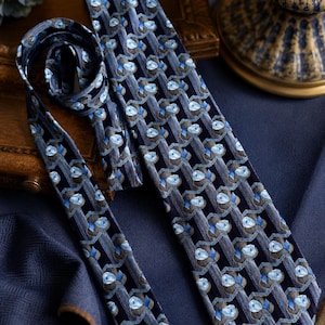 Puede incluir: Una corbata con fondo azul marino oscuro y un patr&oacute;n repetitivo de motivos florales en azul claro y bandas verticales grises. La corbata tiene un extremo puntiagudo y una textura de tela suave, mostrando una disposici&oacute;n geom&eacute;trica.