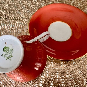 May include: A red teacup and saucer set with gold trim. The teacup has a white interior with a green floral design and the text "EST. 1775 AYNSLEY ENGLAND BONE CHINA 31" on the bottom. The saucer has a white center with a gold rim.
