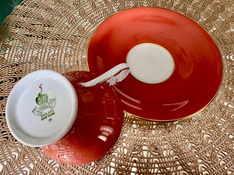 May include: A red teacup and saucer set with gold trim. The teacup has a white interior with a green floral design and the text "EST. 1775 AYNSLEY ENGLAND BONE CHINA 31" on the bottom. The saucer has a white center with a gold rim.