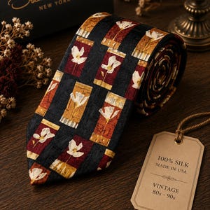 Op de afbeelding: Een opgerolde zwarte zijden stropdas met een bloemenpatroon in rood, goud en wit. Het label luidt "100% Zijde Made in USA Vintage 80s-90s". Het Oscar de la Renta-label is zichtbaar op de achtergrond.