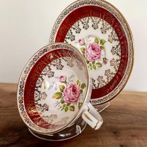 Pode incluir: Um conjunto de ch&aacute; vintage. A ch&aacute;vena e o pires de porcelana branca apresentam um design de borda vermelha e dourada. Uma rosa rosa &eacute; pintada no centro da ch&aacute;vena e do pires. A ch&aacute;vena tem uma pega branca com acabamentos dourados.