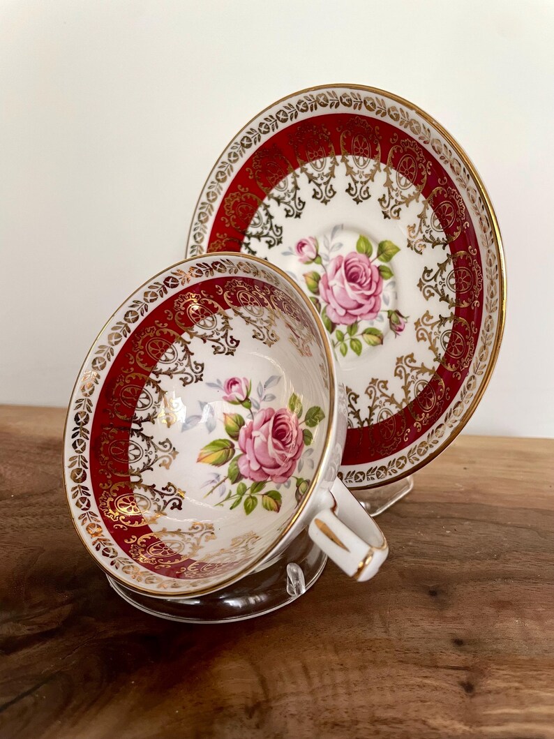 Pode incluir: Um conjunto de ch&aacute; vintage. A ch&aacute;vena e o pires de porcelana branca apresentam um design de borda vermelha e dourada. Uma rosa rosa &eacute; pintada no centro da ch&aacute;vena e do pires. A ch&aacute;vena tem uma pega branca com acabamentos dourados.