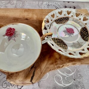 Puede incluir: Un juego de taza y platillo de t&eacute; vintage. La taza y el platillo son blancos con ribetes dorados y presentan un dise&ntilde;o de rosa roja. El platillo tiene un borde en forma de coraz&oacute;n. El juego est&aacute; sobre una superficie de madera.