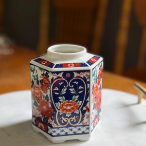 Peut inclure: Un vase hexagonal en c&eacute;ramique avec une base blanche, orn&eacute; d'un motif floral vibrant en rouge, bleu et vert. Le vase pr&eacute;sente un design d&eacute;taill&eacute; avec un fond bleu et des accents floraux rouges, mettant en valeur une esth&eacute;tique classique.