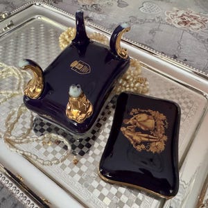 May include: A dark blue porcelain decorative piece with gold accents and a matching small rectangular box with a gold illustration. The pieces are on a silver tray with a checkered pattern and a beaded necklace.