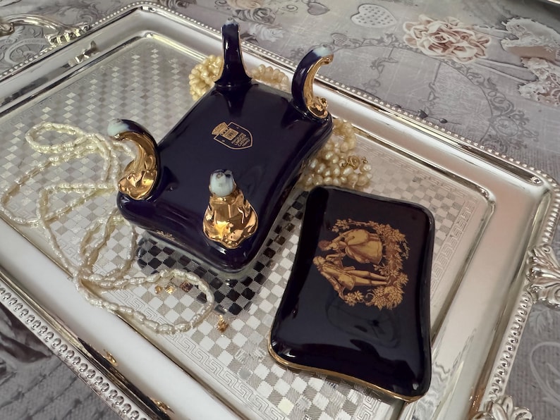 May include: A dark blue porcelain decorative piece with gold accents and a matching small rectangular box with a gold illustration. The pieces are on a silver tray with a checkered pattern and a beaded necklace.