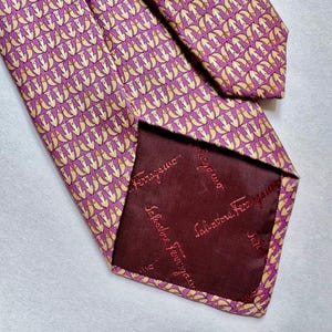 Puede incluir: Una corbata morada con un patr&oacute;n repetitivo de peque&ntilde;os peces amarillos. La parte posterior de la corbata tiene un forro granate oscuro con el texto Salvatore Ferragamo impreso repetidamente en un tono granate m&aacute;s claro.