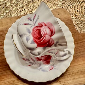 May include: A white teacup and saucer with a floral design featuring a large red rose. The cup and saucer have a gold trim.