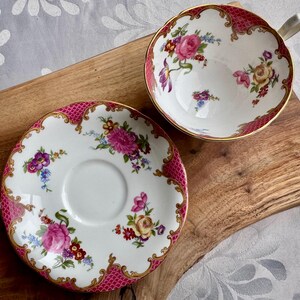 May include: A vintage teacup and saucer set. The white porcelain cup and saucer feature a pink and gold filigree border and are decorated with colorful floral bouquets. The teacup has a handle and the saucer has a central indentation.