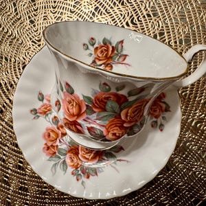 Pode incluir: Um conjunto de ch&aacute;vena e pires branco com um padr&atilde;o floral de rosas laranja e rosa. O conjunto tem uma borda dourada.