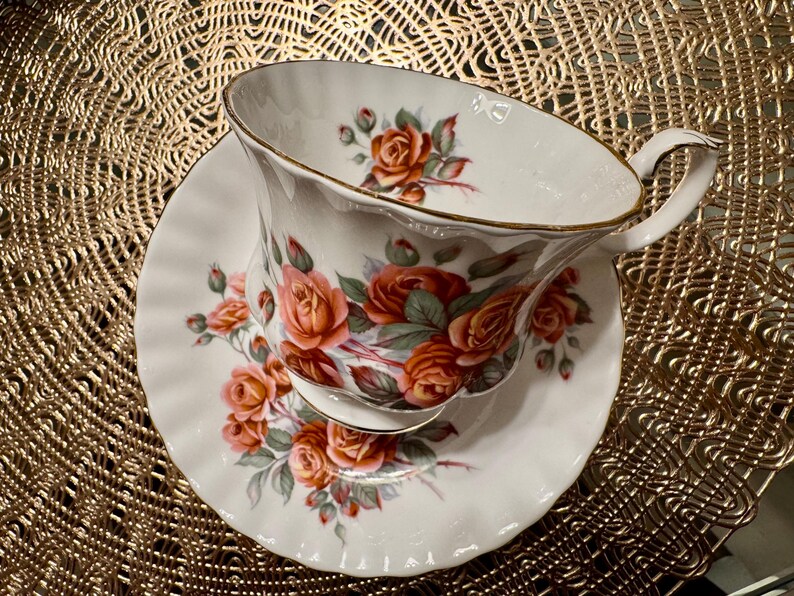 Pode incluir: Um conjunto de ch&aacute;vena e pires branco com um padr&atilde;o floral de rosas laranja e rosa. O conjunto tem uma borda dourada.
