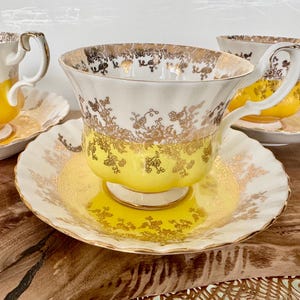 Puede incluir: Un juego de tazas de t&eacute; y platillos vintage. Las tazas y platillos son blancos con una base amarilla y detalles florales dorados. Los bordes y las asas tambi&eacute;n est&aacute;n adornados en dorado. Las tazas de t&eacute; est&aacute;n sobre una superficie de madera.