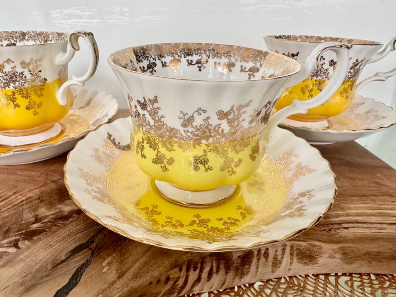 Puede incluir: Un juego de tazas de t&eacute; y platillos vintage. Las tazas y platillos son blancos con una base amarilla y detalles florales dorados. Los bordes y las asas tambi&eacute;n est&aacute;n adornados en dorado. Las tazas de t&eacute; est&aacute;n sobre una superficie de madera.