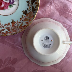 May include: A fine bone china teacup and saucer set. The saucer features a light blue rim with a floral pattern and gold accents. The teacup is inverted, showing a black maker's mark that reads BY APPOINTMENT TO HER MAJESTY THE QUEEN PARAGON.
