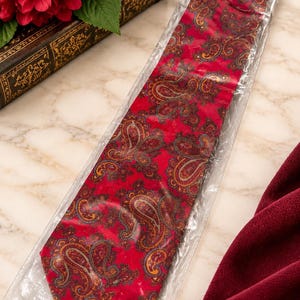 May include: A red necktie featuring a repeating paisley pattern in shades of gold, blue, and dark brown. The necktie is encased in a clear plastic protective sleeve, showcasing its pointed tip and smooth, woven fabric construction.