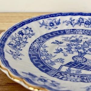 May include: A white and blue porcelain plate with a floral design. The plate features a blue floral pattern around the rim and a central design of flowers in a vase. The rim is scalloped and trimmed with gold.