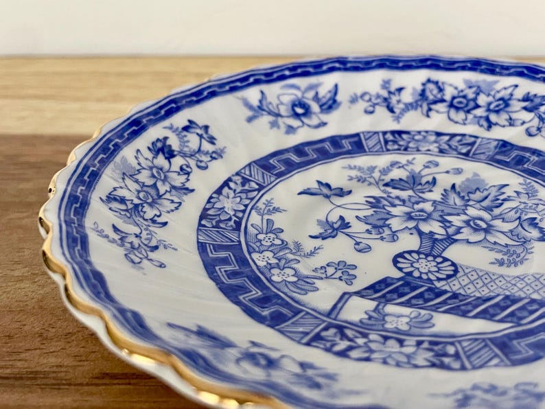 May include: A white and blue porcelain plate with a floral design. The plate features a blue floral pattern around the rim and a central design of flowers in a vase. The rim is scalloped and trimmed with gold.