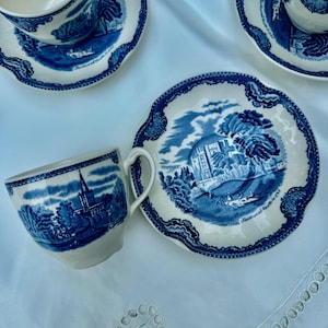 Puede incluir: Un juego de tazas y platillos azules y blancos vintage. Cada pieza presenta una escena de paisaje detallada con una iglesia o castillo, y bordes festoneados decorativos. El juego est&aacute; dispuesto sobre un mantel blanco.