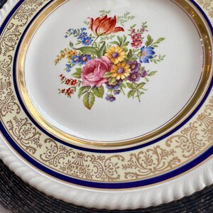 May include: A white ceramic plate with a floral bouquet design in the center. The bouquet features a red tulip, pink rose, and various other colorful flowers. The plate has a gold rim and a blue border with a decorative pattern.