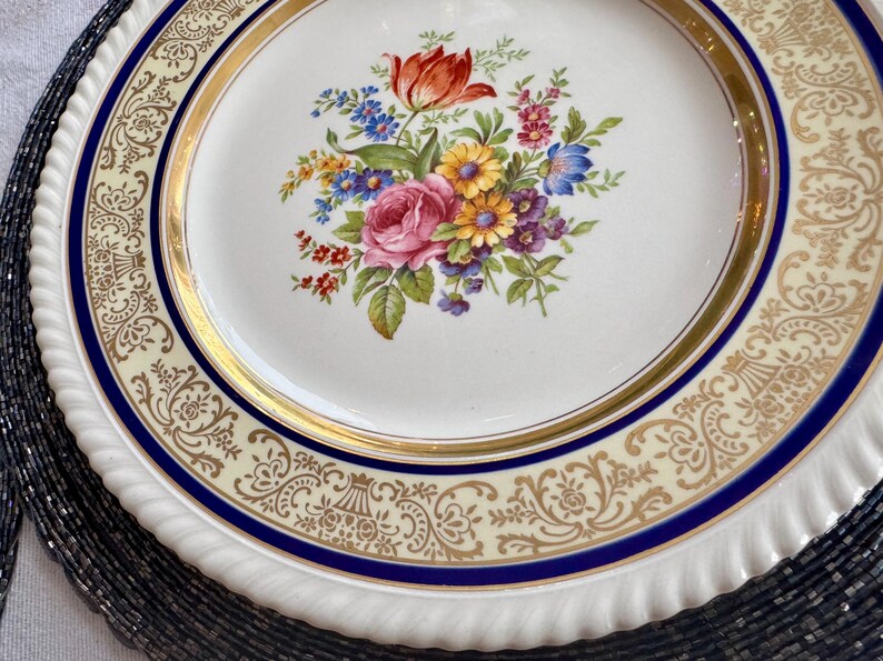May include: A white ceramic plate with a floral bouquet design in the center. The bouquet features a red tulip, pink rose, and various other colorful flowers. The plate has a gold rim and a blue border with a decorative pattern.