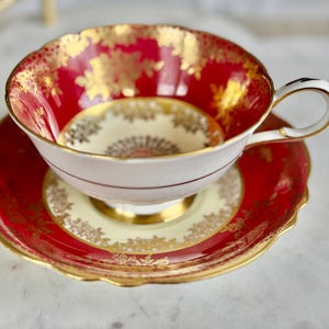 May include: A vintage teacup and saucer set. The teacup features a red interior with gold floral accents and a white exterior. The saucer is red with gold trim. The set is designed with a scalloped edge and a gold handle.
