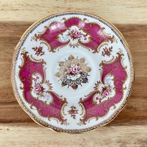 May include: A vintage teacup saucer with a white center and a vibrant pink border. The saucer features intricate gold filigree and floral designs, with a scalloped gold rim. The overall aesthetic is ornate and elegant.