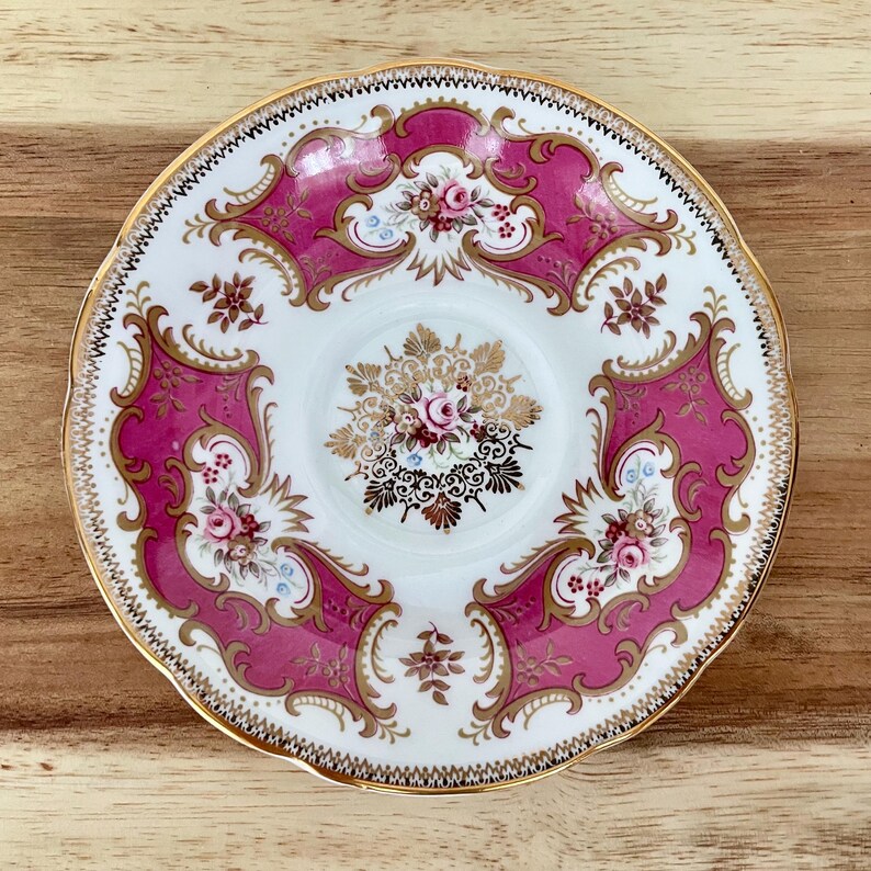May include: A vintage teacup saucer with a white center and a vibrant pink border. The saucer features intricate gold filigree and floral designs, with a scalloped gold rim. The overall aesthetic is ornate and elegant.