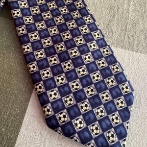 May include: A necktie featuring a repeating checkered pattern of dark blue squares and light squares. Each light square contains a graphic illustration of a black and white soccer ball. The tie has a pointed end and a smooth, woven fabric texture.