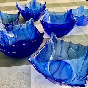 Pu&ograve; includere: Una collezione di sette ciotole in vetro blu intenso con un motivo strutturato simile a una foglia sull'esterno. Ogni ciotola presenta una forma quadrata con bordi irregolari e ondulati che imitano l'aspetto di una foglia naturale.