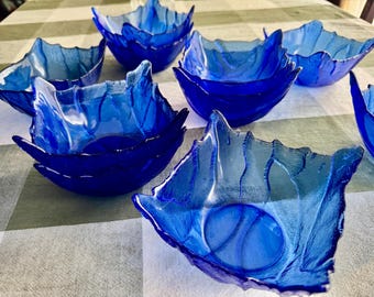 May include: A collection of seven deep blue glass bowls with a textured, leaf-like pattern on the exterior. Each bowl features a square shape with irregular, wavy edges that mimic the appearance of a natural leaf, creating a translucent effect.