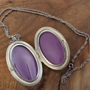 Puede incluir: Collar con colgante ovalado plateado, abierto para revelar un interior p&uacute;rpura. El colgante cuelga de una delicada cadena. El colgante est&aacute; sobre una superficie de madera.