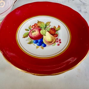 May include: A red and white porcelain saucer with a gold rim. The center of the saucer features a white circle with a hand-painted design of fruit, including an apple, pear, grapes, and strawberries.