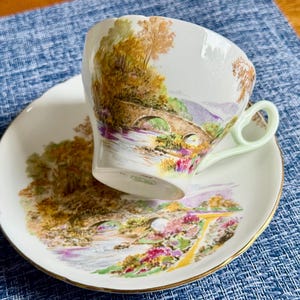 May include: A white porcelain teacup and saucer set featuring a colorful landscape illustration of a stone bridge over a river. The cup has a pale green handle, and both pieces are decorated with autumn-toned trees, foliage, and a gold-trimmed rim.