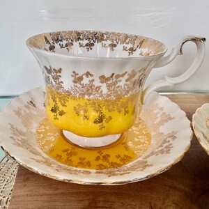 Puede incluir: Juego de taza y platillo de t&eacute; vintage. La taza y el platillo son blancos con dise&ntilde;os florales dorados y una banda amarilla. La taza tiene un asa curvada. El juego est&aacute; sobre una superficie de madera.