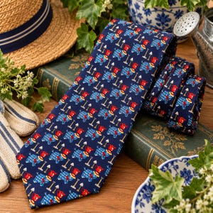 May include: A navy blue necktie featuring a repeating pattern of small red watering cans and white garden rakes. The fabric has a smooth finish, and the design is oriented vertically along the length of the tie, showcasing a consistent garden-themed motif.