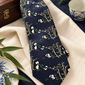 May include: A dark navy blue necktie featuring a repeating pattern of black and white pandas eating green bamboo stalks. The fabric has a smooth finish and the design is distributed evenly across the entire length of the necktie.