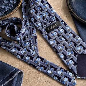 Puede incluir: Una corbata azul marino con un patr&oacute;n repetitivo de formas florales en azul claro y gris dispuestas en columnas verticales. La corbata incluye una etiqueta negra con el texto blanco PROFILO STUDIO cosido en la parte posterior.