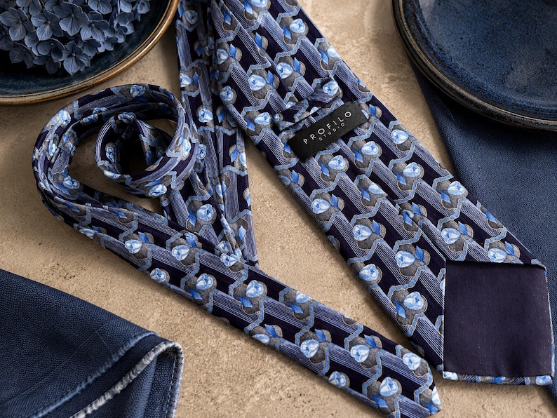Puede incluir: Una corbata azul marino con un patr&oacute;n repetitivo de formas florales en azul claro y gris dispuestas en columnas verticales. La corbata incluye una etiqueta negra con el texto blanco PROFILO STUDIO cosido en la parte posterior.