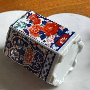 Peut inclure: Un vase en c&eacute;ramique blanc de forme rectangulaire. Le vase est d&eacute;cor&eacute; de motifs floraux rouges et oranges, de feuilles bleues et d'accents dor&eacute;s. Le motif est pos&eacute; sur un fond bleu fonc&eacute;.