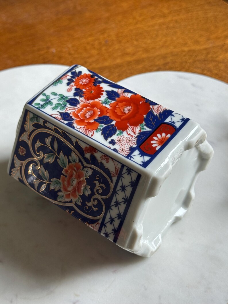 Peut inclure: Un vase en c&eacute;ramique blanc de forme rectangulaire. Le vase est d&eacute;cor&eacute; de motifs floraux rouges et oranges, de feuilles bleues et d'accents dor&eacute;s. Le motif est pos&eacute; sur un fond bleu fonc&eacute;.